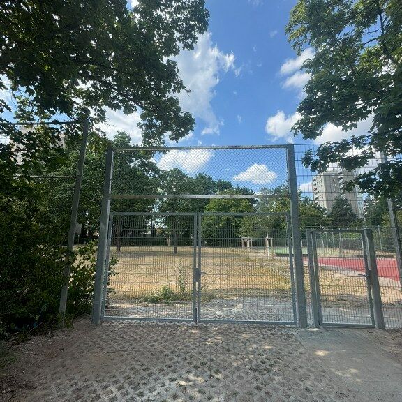 Der Hartplatz nach der Zaunschließung. Man sieht das Eingangstor, den Bolzplatz und den neuen Zaun außen herum.